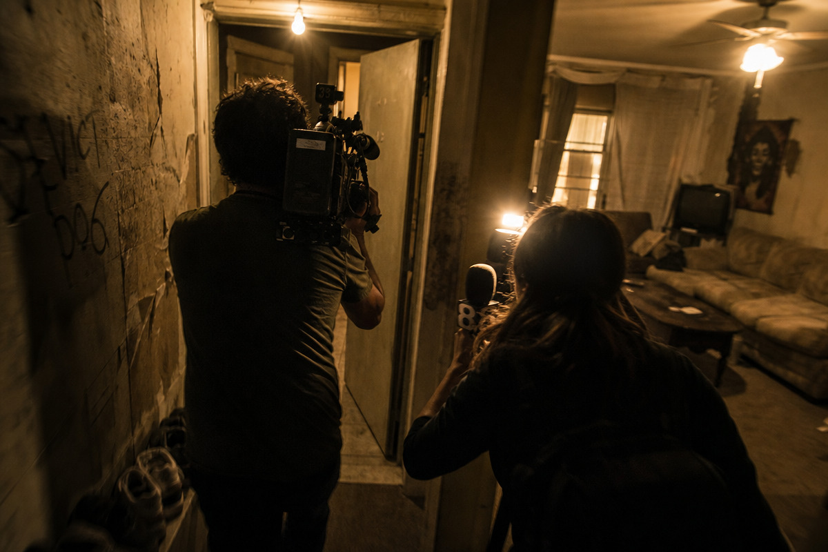 A camera operator and reporter move through a cramped apartment corridor toward a half-open door.