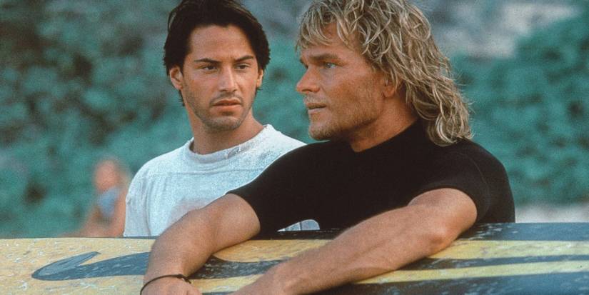 Keanu Reeves and Patrick Swayze outdoors with a surfboard in Point Break.